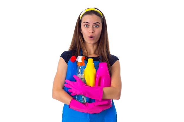 Giovane donna sorpresa in maglietta blu e grembiule con guanti rosa che tengono detersivi in studio
