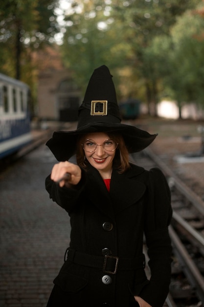 Giovane donna sorride sorniona e vestita con cappello da streghe cappotto nero e occhiali con bacchetta magica in mano Cornice verticale