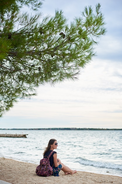 Giovane donna sorridente attraente con lo zaino variopinto che si siede da solo sulla spiaggia sabbiosa dell'oceano su chiari acqua e cielo blu luminosi
