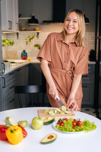 Giovane donna sorridente che affetta le zucchine per preparare insalata vegetariana al tavolo su tavola di legno in cucina con interni moderni, guardando la telecamera. Concetto di alimentazione sana.