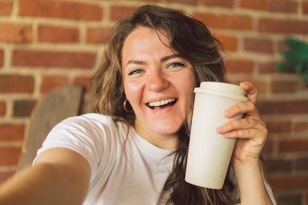 Giovane donna sorridente che beve caffè e guarda a porte chiuse e prende un concetto di stile di vita selfie