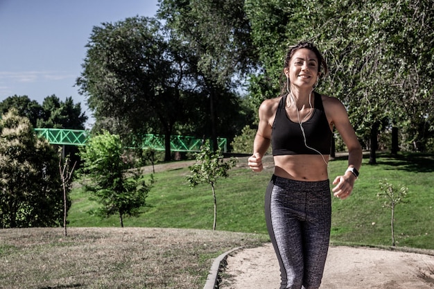 Giovane donna sorridente che corre attraverso un parco in una giornata di sole mentre ascolti musica con le cuffie