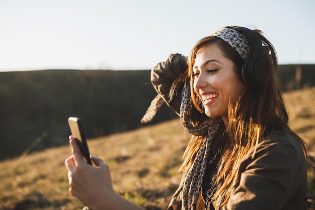 Giovane donna sorridente che effettua una videochiamata tramite smartphone mentre si siede per terra in una giornata di sole e ascolta musica.