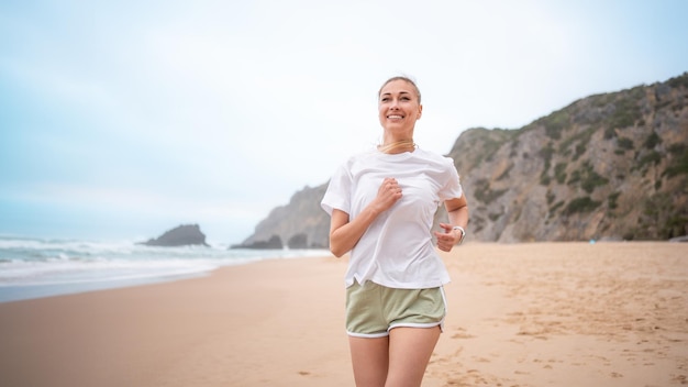 Giovane donna sorridente che fa jogging sulla spiaggia di sabbia lungo le onde del mare