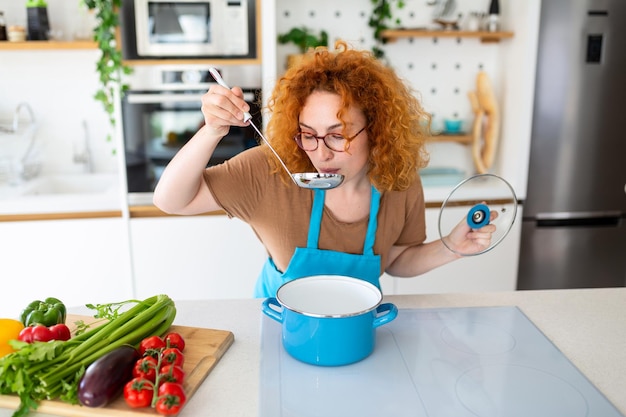 Giovane donna sorridente che gode del gusto e dell'olfatto mentre cucina il pranzo in cucina