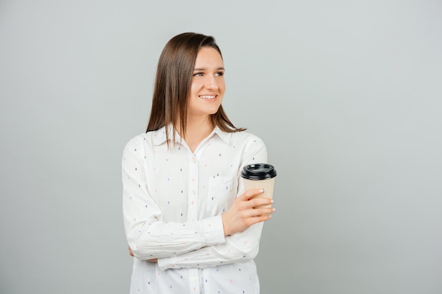 Giovane donna sorridente che guarda lontano è in possesso di una tazza di caffè da asporto Studio girato su sfondo grigio