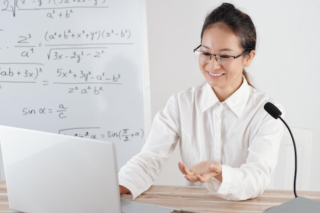 Giovane donna sorridente che insegna a distanza da casa lavagna con formule matematiche in background