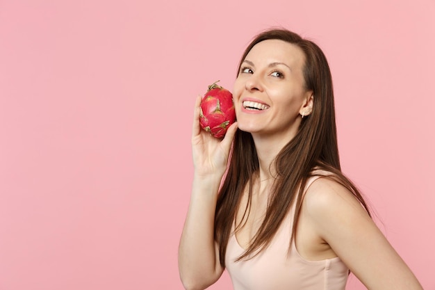 Giovane donna sorridente che osserva in su tenendo in mano pitahaya maturo fresco, frutta del drago isolata su fondo rosa pastello della parete in studio. Lo stile di vita vivido delle persone si rilassa il concetto di vacanza. Mock up spazio di copia.