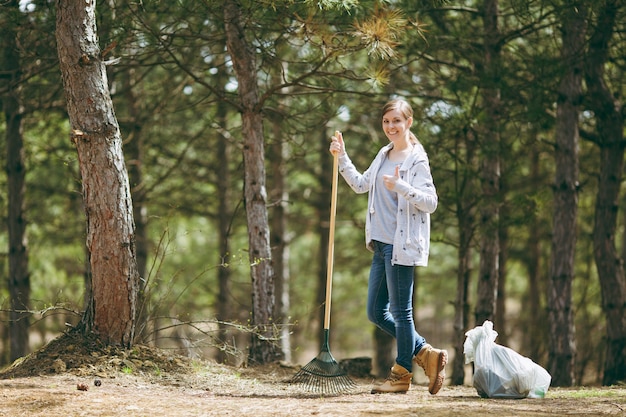 Giovane donna sorridente che pulisce con il rastrello per la raccolta dei rifiuti e mostra il pollice vicino ai sacchetti della spazzatura nel parco. Problema di inquinamento ambientale