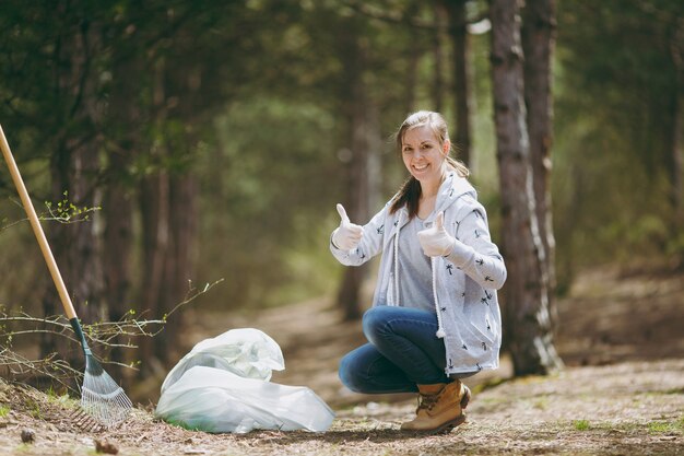 Giovane donna sorridente che pulisce la spazzatura nei sacchetti della spazzatura che mostra i pollici in su nel parco o nella foresta