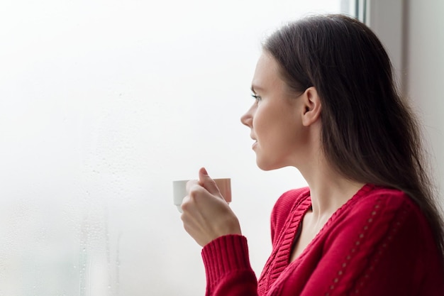 Giovane donna sorridente che si gode una donna di caffè profumato in un caldo maglione lavorato a maglia vicino allo spazio della copia della finestra Stile di vita a casa stagione autunno inverno