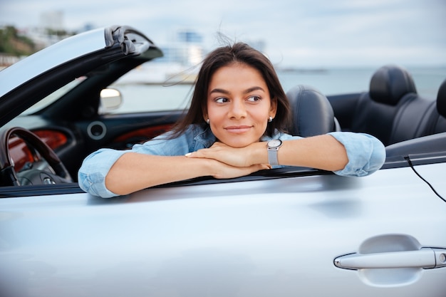 Giovane donna sorridente che si siede all'interno della sua automobile convertibile sulla spiaggia