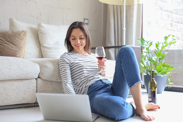 Giovane donna sorridente che si siede sul pavimento con il computer portatile e che chiacchiera con gli amici che bevono vino