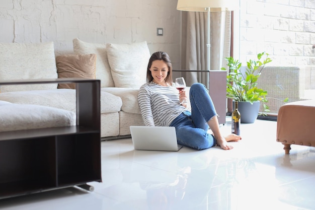 Giovane donna sorridente che si siede sul pavimento con il computer portatile e che chiacchiera con gli amici che bevono vino