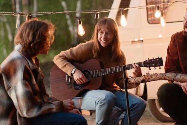 Giovane donna sorridente che suona la chitarra e canta canzoni insieme ai suoi amici durante un picnic