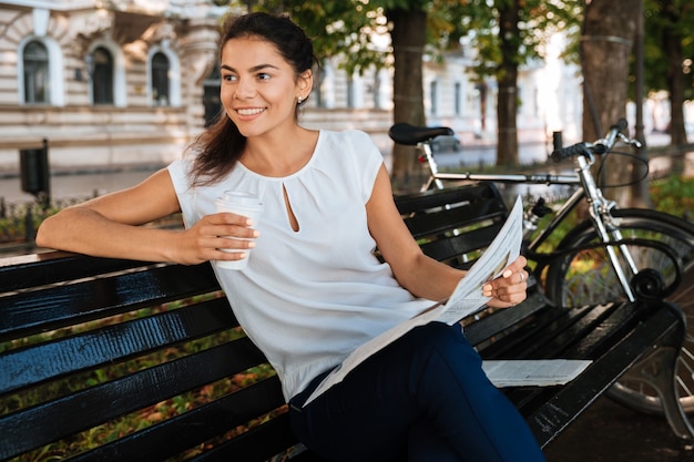 Giovane donna sorridente che tiene il giornale mentre era seduto sulla panchina con una tazza di caffè