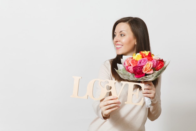 Giovane donna sorridente che tiene la parola in legno amore, bouquet di bellissime rose fiori isolati su sfondo bianco. Copia spazio per la pubblicità. Concetto di San Valentino o Giornata internazionale della donna.