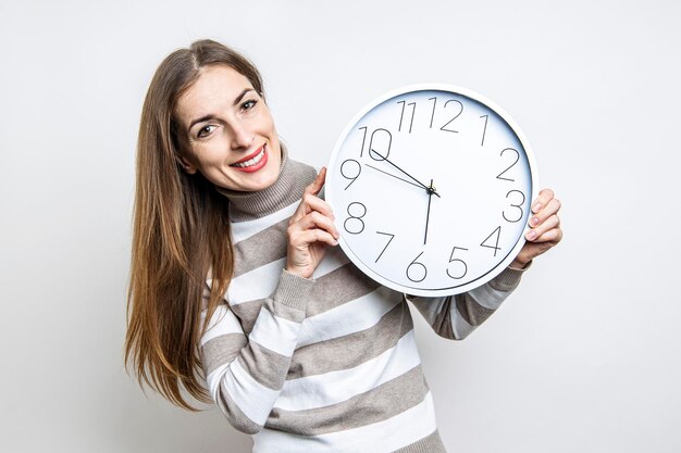 Giovane donna sorridente che tiene orologio da parete bianco su sfondo chiaro