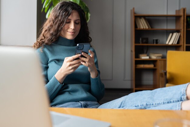 Giovane donna sorridente che tiene smartphone in chat online mentre ci si rilassa nell'accogliente ufficio a casa