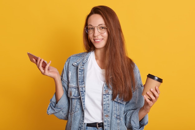 giovane donna sorridente che tiene telefono mobile e bere portare via caffè, navigare internatore o chattare con gli amici