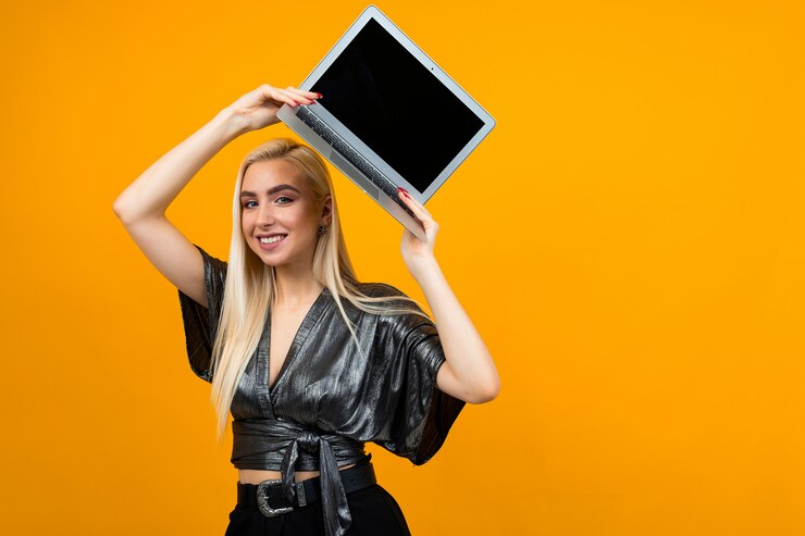 Foto giovane donna sorridente che tiene un computer portatile con uno schermo in bianco per inserire una pagina web sulla parete gialla