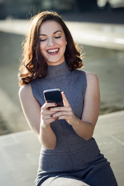 Giovane donna sorridente che usa il cellulare mentre è seduta fuori da un edificio moderno
