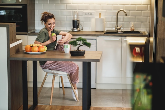 Giovane donna sorridente che usa il suo smartphone mentre mangia la mela al mattino a casa.