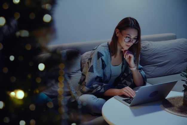 Giovane donna sorridente che utilizza il laptop di notte o una videochiamata serale seduta sul divano di casa sullo sfondo dell'albero di Natale e delle luci