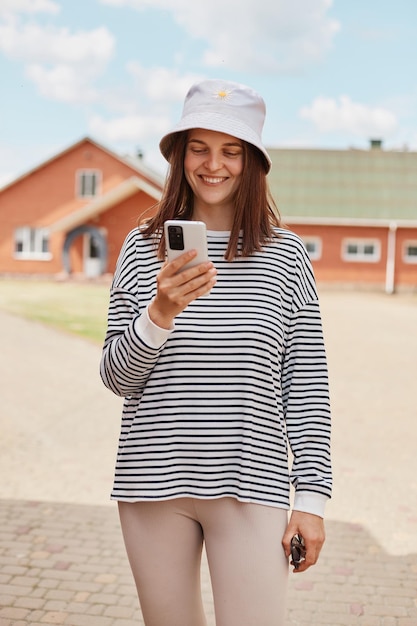 Giovane donna sorridente che utilizza lo smartphone in piedi sulla strada vicino alle case che indossano la camicia a righe utilizzando il telefono cellulare controllando i social network mentre si cammina all'aperto