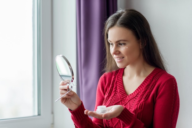 Giovane donna sorridente con crema idratante per il viso vicino agli occhi crema per il viso femminile in piedi vicino alla finestra con specchio Concetto di cura della pelle e della gioventù