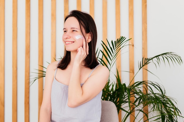 Giovane donna sorridente con gli occhi chiusi che applica crema sul viso a casa concetto di cura della pelle di routine di bellezza