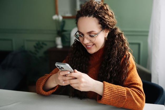 Giovane donna sorridente con gli occhiali in possesso di uno smartphone utilizzando la tecnologia moderna del cellulare guardando il cellulare controllando le app del telefono cellulare mandando SMS e navigando in Internet per lo shopping seduto a casa