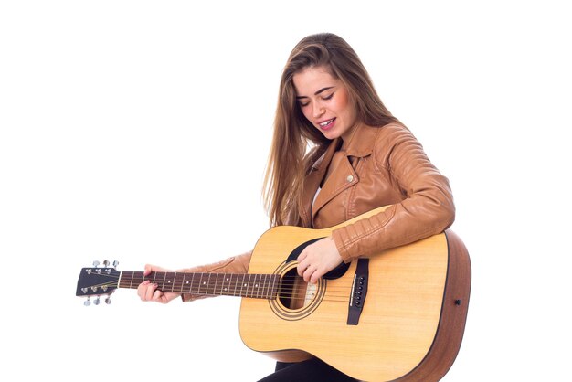 Giovane donna sorridente con i capelli lunghi in giacca marrone che suona la chitarra su sfondo bianco in studio