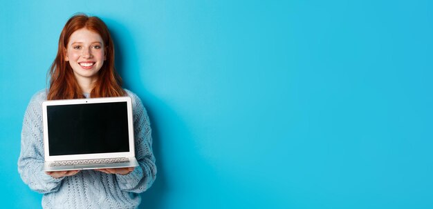 Giovane donna sorridente con i capelli rossi e le lentiggini che mostra lo schermo del computer che tiene il portatile e dimostra