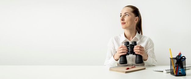 Giovane donna sorridente con il binocolo seduto alla scrivania dell'ufficio su sfondo bianco Banner