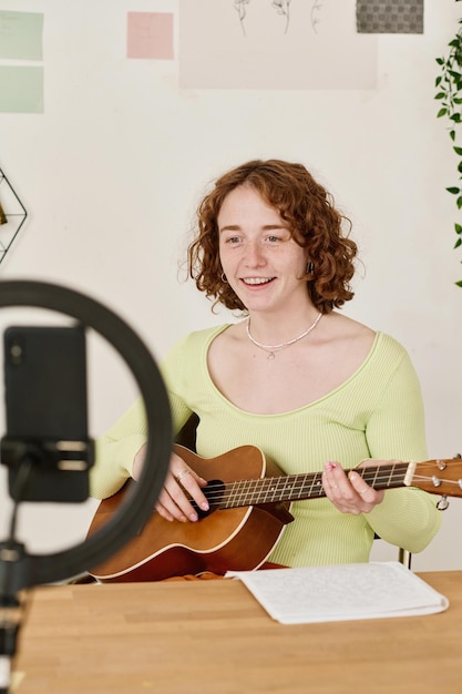 Giovane donna sorridente con la chitarra acustica che fa lezione video per il pubblico online