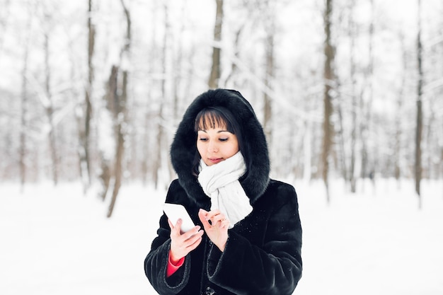 Giovane donna sorridente con smart phone e paesaggio invernale e fiocchi di neve sullo sfondo.