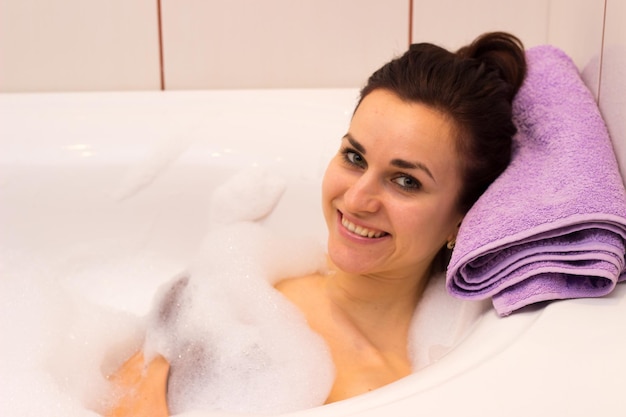 Giovane donna sorridente con un fagotto sulla testa sdraiata nella vasca da bagno piena di schiuma