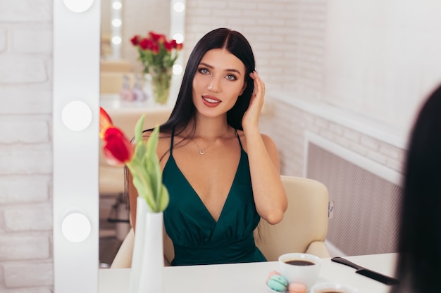 Giovane donna sorridente del brunette con la tazza di caffè che esamina lo specchio nel salone di parrucchiere