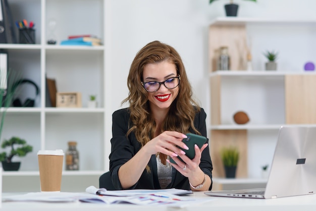 Giovane donna sorridente di affari in vestiti alla moda che si siedono nell'ufficio moderno accogliente e che passano in rassegna qualcosa sullo smartphone.