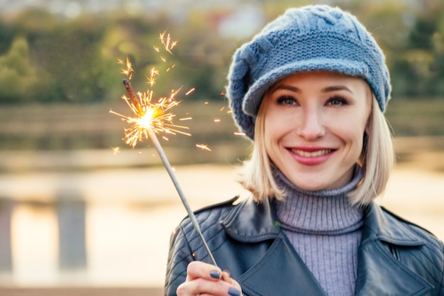 Giovane donna sorridente felice bionda in cappotto beige e cappello caldo con luci del Bengala nel suo ritratto all'aperto del primo piano delle mani nel parco autunnale. Freddo fuori. Vestiti primaverili invernali, Natale, concetto di Capodanno