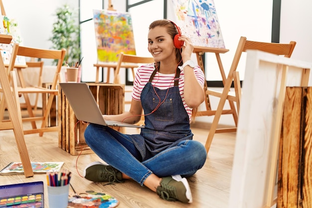 Giovane donna sorridente fiducioso utilizzando il computer portatile e le cuffie in studio d'arte