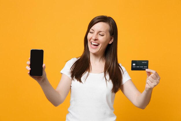 Giovane donna sorridente in abiti casual bianchi che tiene il telefono cellulare con una carta di credito vuota con schermo vuoto isolato su sfondo giallo arancione in studio. Concetto di stile di vita delle persone. Mock up spazio di copia.