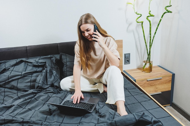 Giovane donna sorridente in abiti casual che sorride e lavora a distanza seduta sul letto con il computer portatile durante il lavoro a distanza Lavoro freelance Lavoro da casa