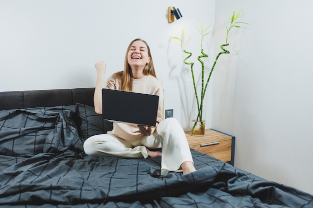 Giovane donna sorridente in abiti casual che sorride e lavora a distanza seduta sul letto con il computer portatile durante il lavoro a distanza Lavoro freelance Lavoro da casa