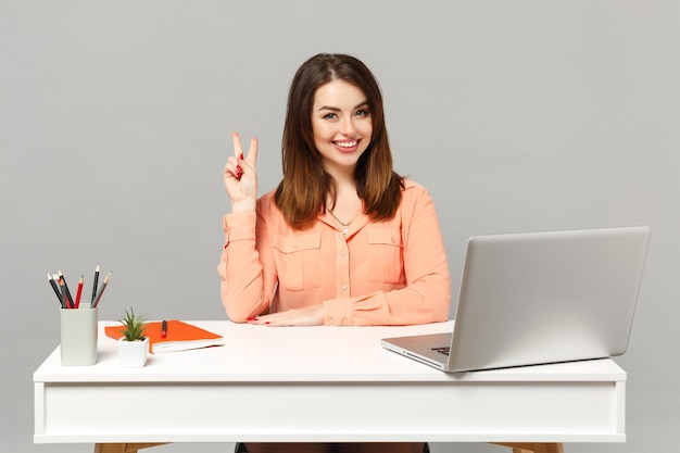 Giovane donna sorridente in abiti casual pastello che mostrano il segno della vittoria sedersi, lavorare alla scrivania bianca con un computer portatile isolato su sfondo grigio. Concetto di stile di vita di carriera aziendale di successo. Mock up spazio di copia.