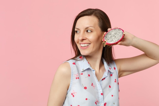 Giovane donna sorridente in abiti estivi che guarda da parte tiene metà del frutto fresco maturo del drago pitahaya isolato su sfondo rosa pastello. Lo stile di vita vivido delle persone si rilassa il concetto di vacanza. Mock up spazio di copia.