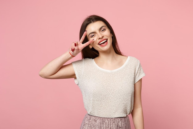 Giovane donna sorridente in abiti leggeri casual in posa isolata su sfondo rosa pastello, ritratto in studio. Persone sincere emozioni concetto di stile di vita. Mock up spazio di copia. Mostra il segno della vittoria.