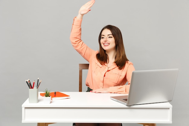 Giovane donna sorridente in abiti pastello che saluta e saluta con la mano mentre nota qualcuno, lavora alla scrivania con un laptop isolato su sfondo grigio. Concetto di carriera aziendale di successo. Mock up spazio di copia.