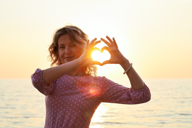 Giovane donna sorridente in abito rosa con le dita a forma di cuore sta al tramonto